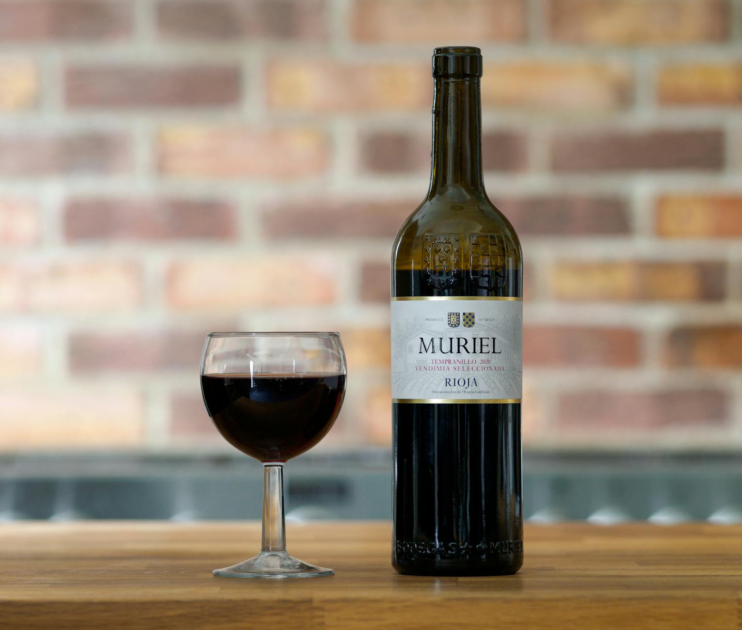 Elegant still life of Muriel Rioja wine bottle with glass on a wooden table against a brick wall backdrop.