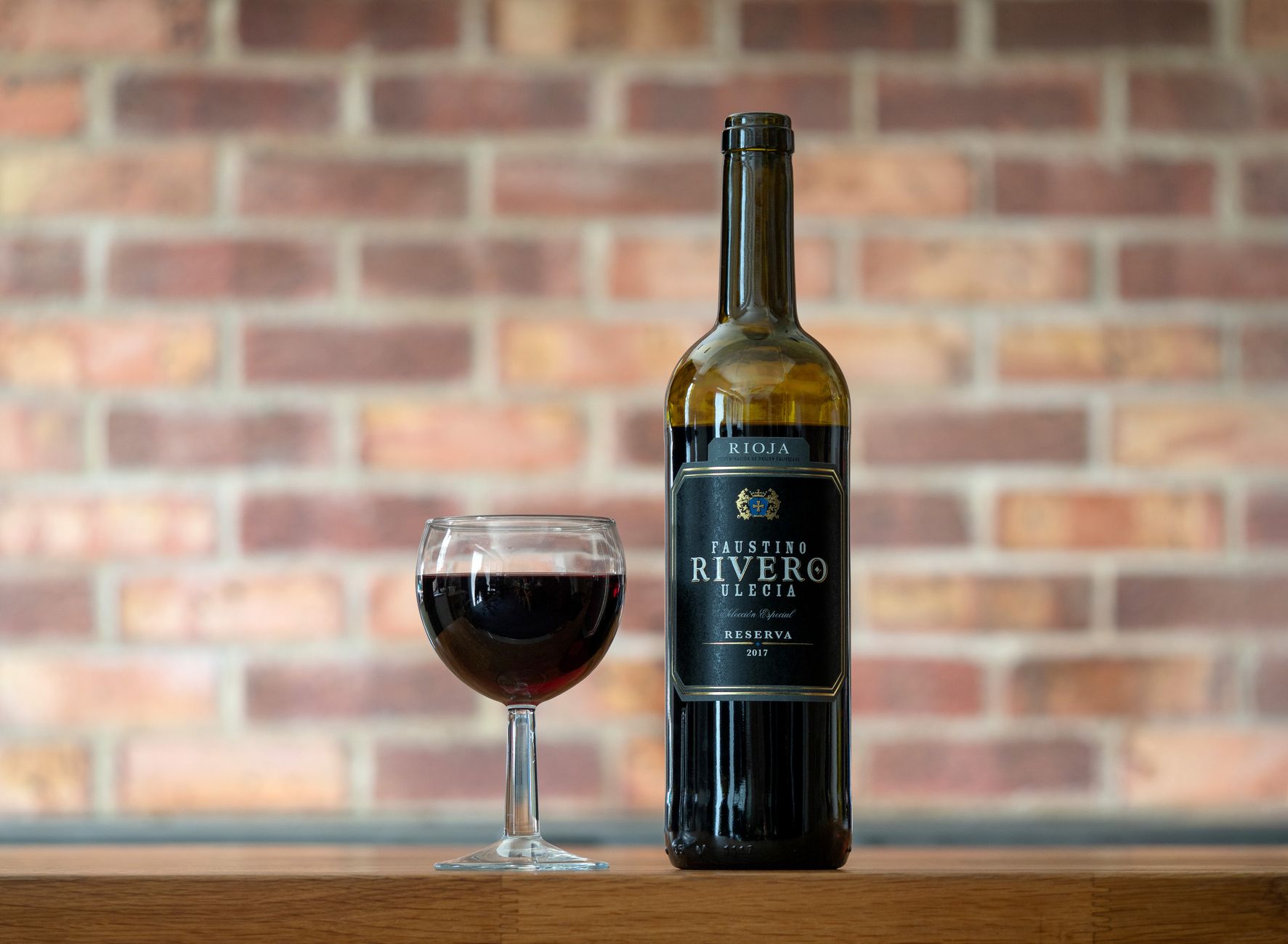 Faustino Rivero red wine bottle and glass set against a brick wall in a cozy indoor setting.
