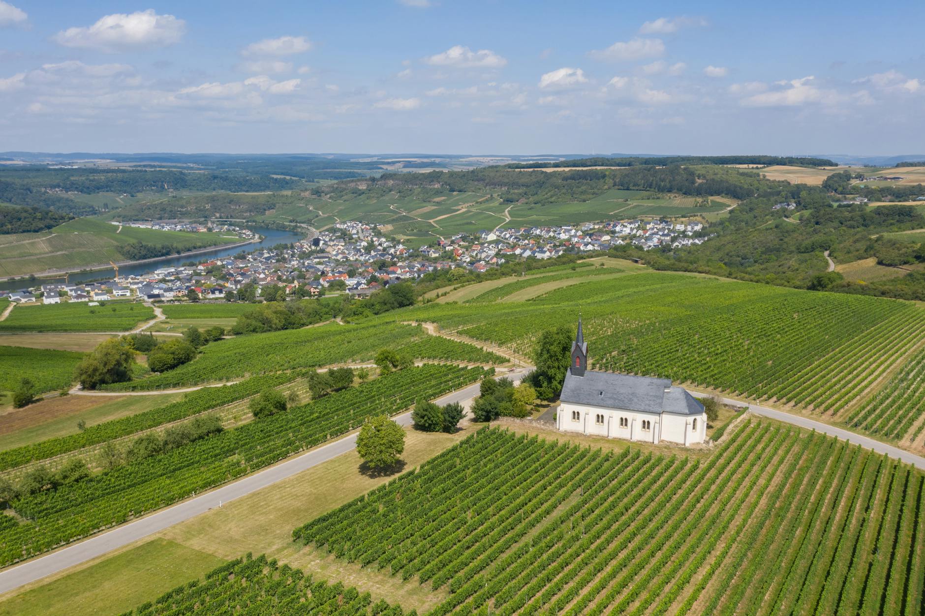Drone view of peaceful countryside with small settlement and church surrounded by green plantations and trees with river on background in summer sunny day with blue sky