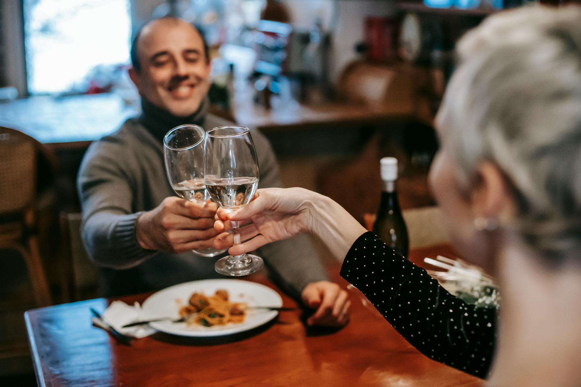 Cheerful adult couple in casual outfits clinking glasses with white wine while having dinner in restaurant