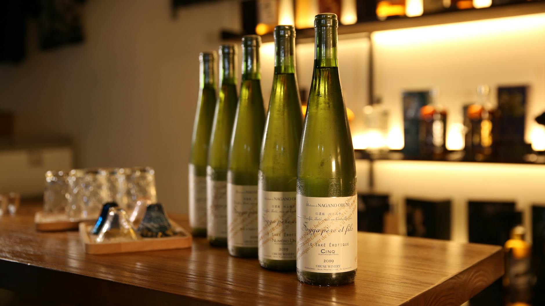 Line of wine bottles on a wooden table with a blurred bar background, creating a warm ambiance.