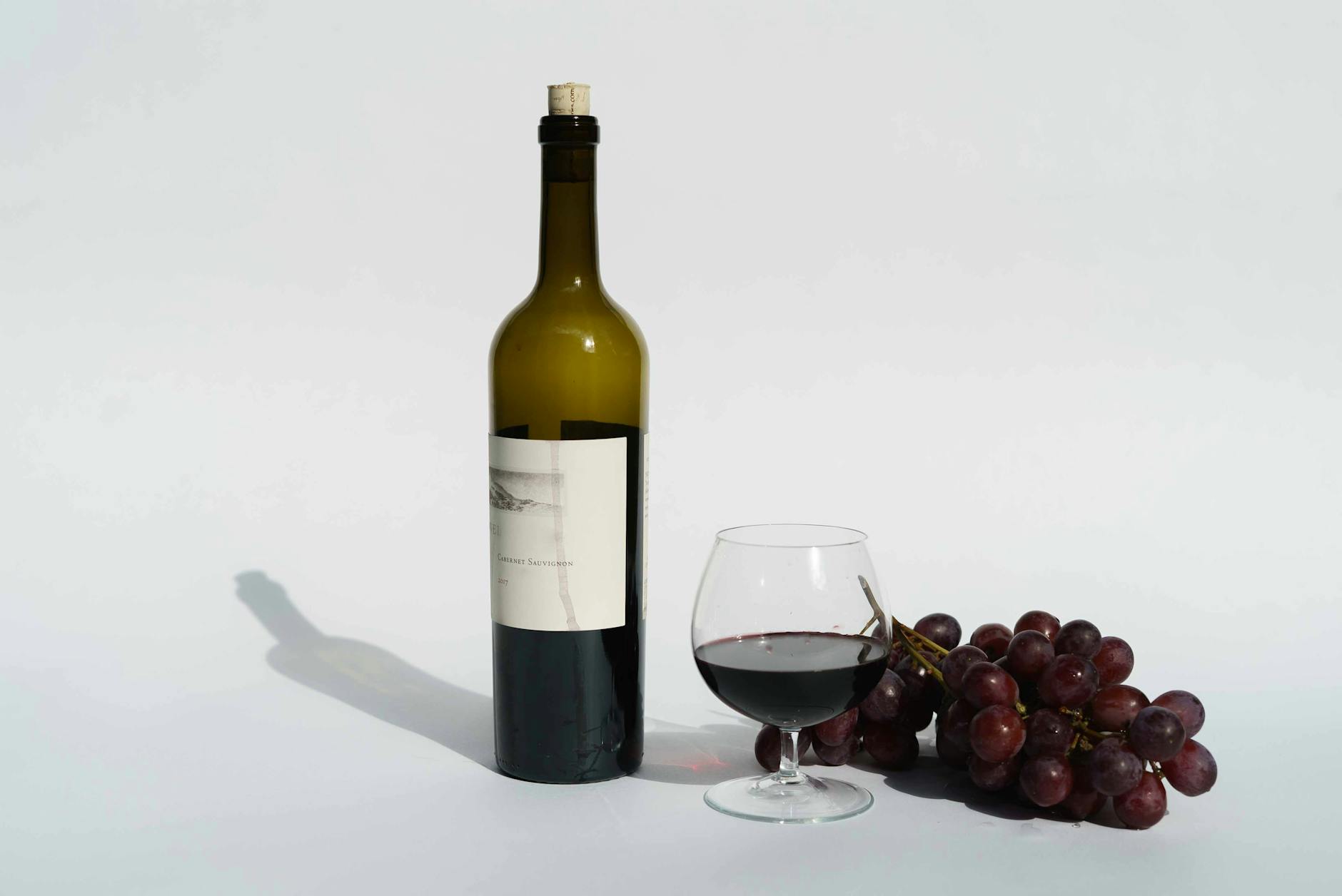 Elegant still life of Cabernet Sauvignon bottle, glass, and grapes on a white background.